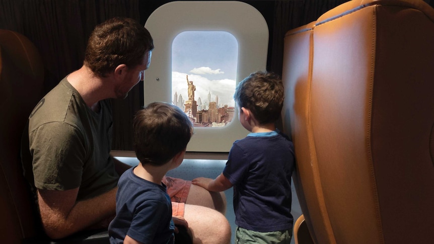 A man and two young boys look out a pretend window at the Statue of Liberty inside an interactive display inside an aircraft