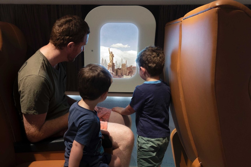 A man and two young boys look out a pretend window at the Statue of Liberty inside an interactive display inside an aircraft