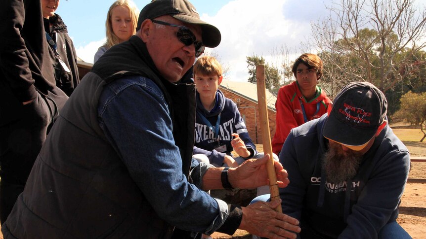 Students watch on as an Aboriginal elder starts a fire using a stone and a stick.