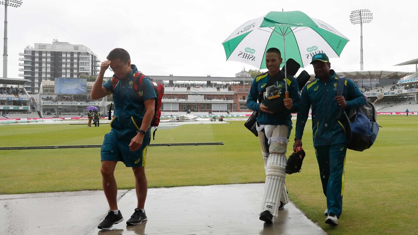 Usman Khawaja walks alongside a coach beneath an umbrella as the rain comes down at Lord's Cricket Ground