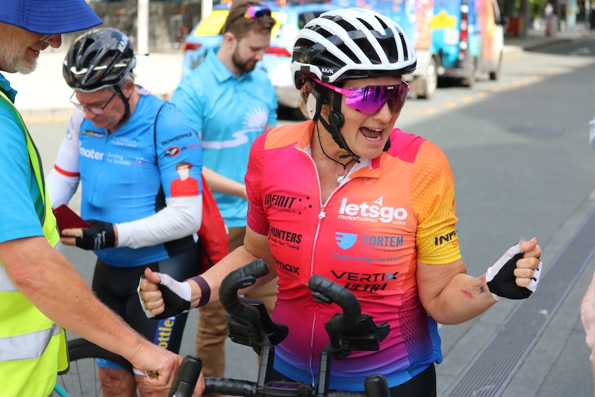 A woman wearing in a cycling suit, wearing sunglasses celebrates with arms out.