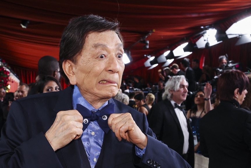 An elderly man with black hair makes a comic expression as he adjusts a bow tie with googly eyes on a Hollywood red carpet.