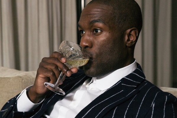 Yahya Abdul-Mateen drinks out of a glass and wears a striped black suit