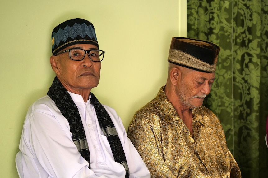 Two Cocos Malay men wearing Taqiyah cap against a light green wall. One man looks at the camera. 