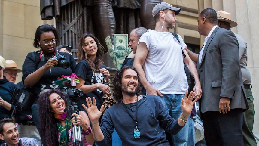 Actor Russell Brand kneels on concrete statute with smiling protestors behind him taking photos.