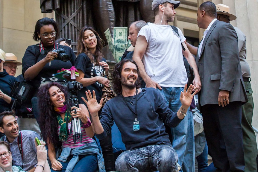 Actor Russell Brand kneels on concrete statute with smiling protestors behind him taking photos.