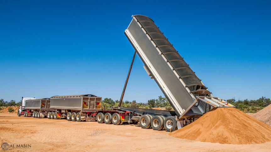 A road train tipping sand to the ground from the back trailer. The trailers are silver.