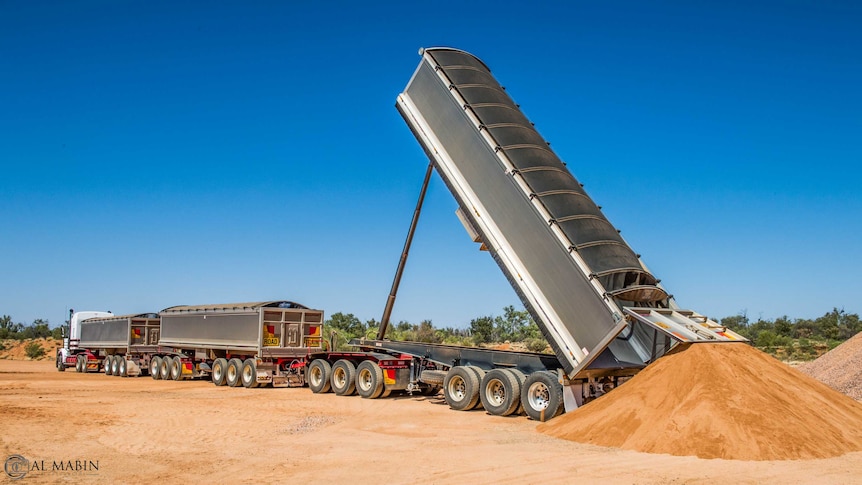 A road train tipping sand to the ground from the back trailer. The trailers are silver.