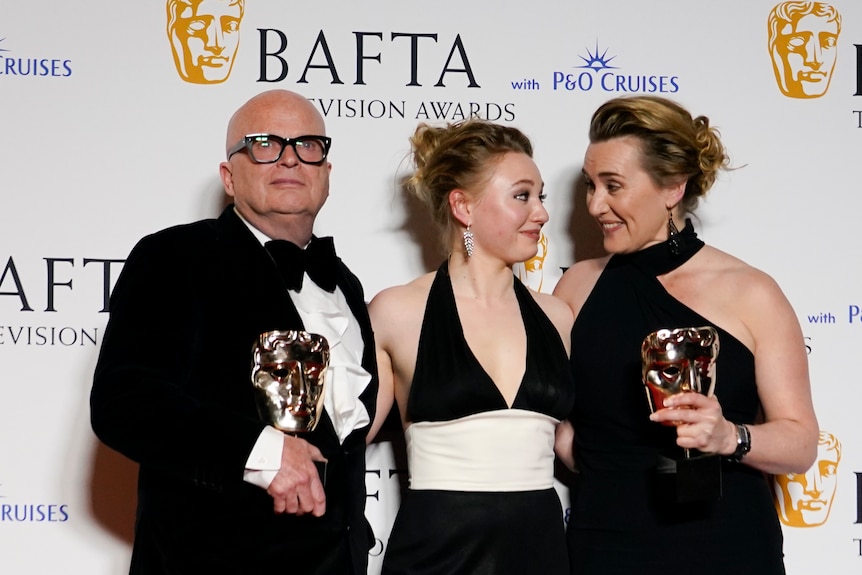 Dominic Savage, Mia Threapleton and Kate Winslet pose while holding BAFTA awards. Threapleton and WInslet share a glance. 