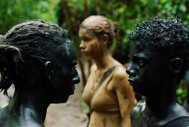 A dreadlocked teen boy covered in black paint stands closely and shouts at younger boy also in black paint near others in jungle