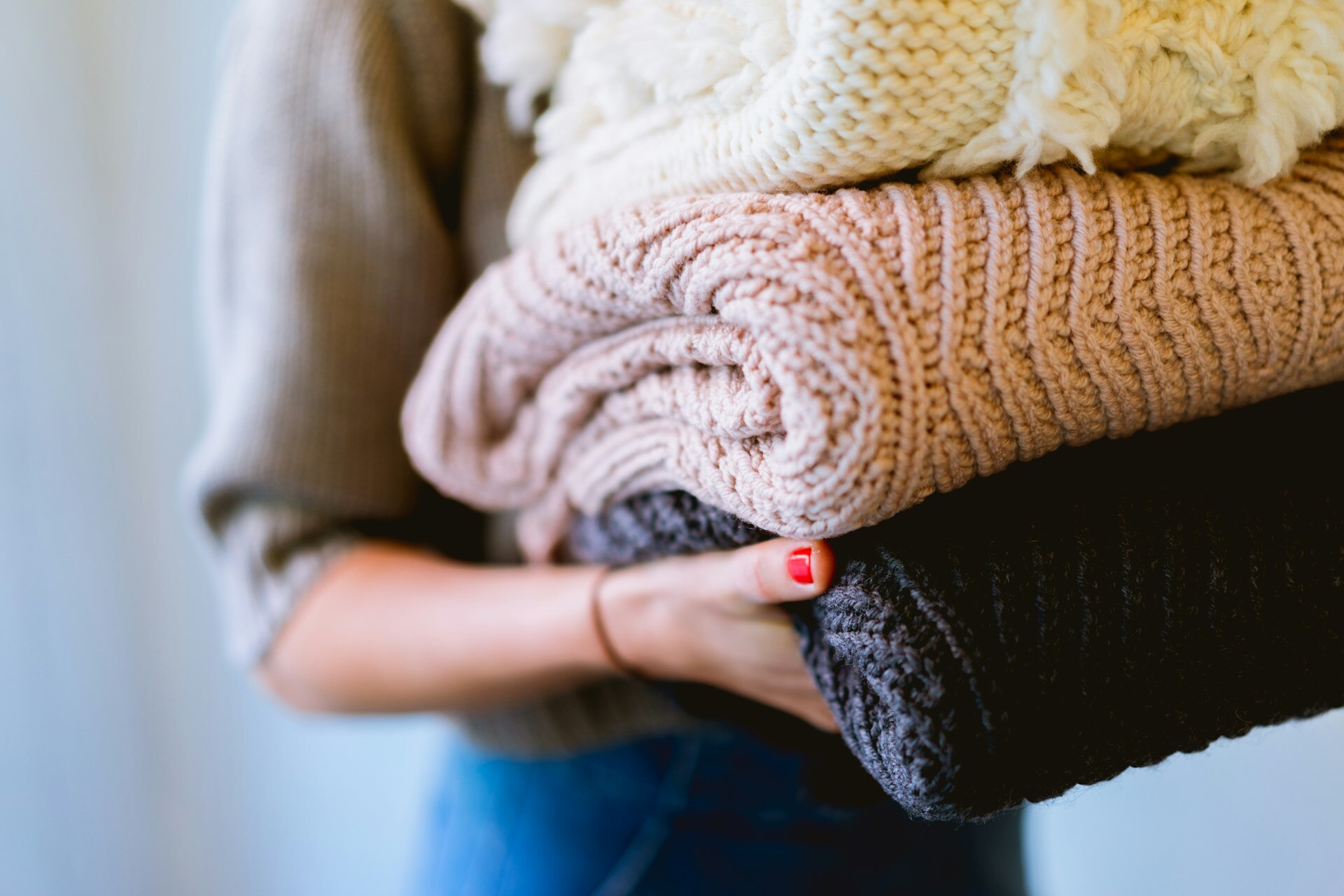 A close up of a stack of folded, thick woolen jumpers, held in the arms of a woman.