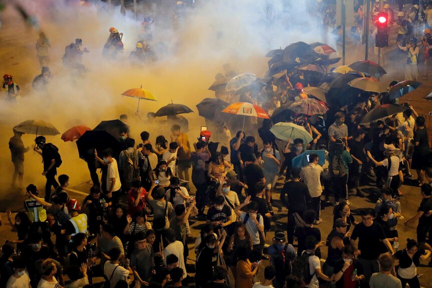 Police fire tear gas into a crowd as dozens of people hold umbrellas.