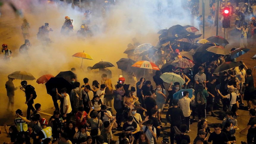 Police fire tear gas into a crowd as dozens of people hold umbrellas.