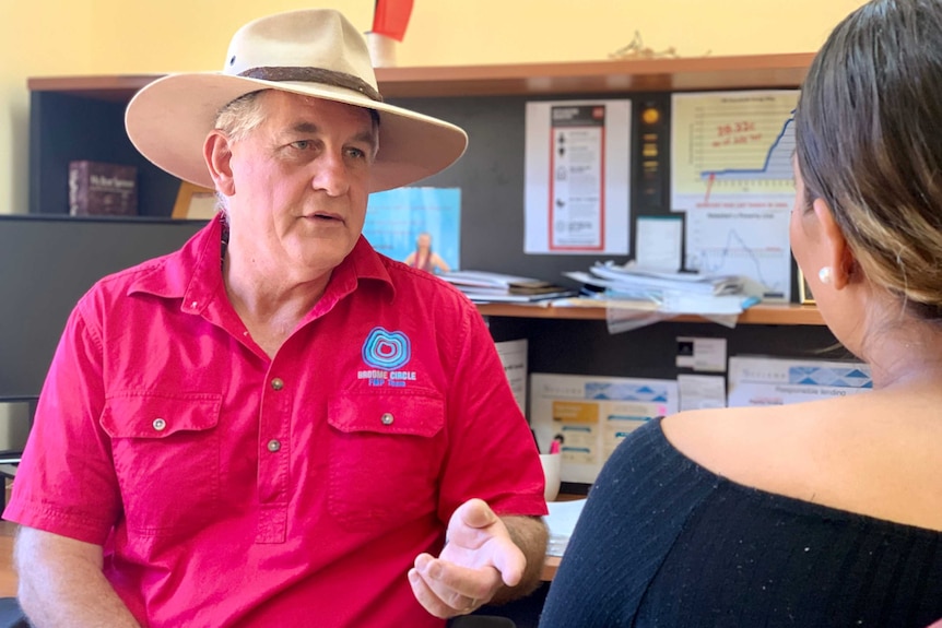 A man wearing a hat sits in an office speaking with a journalist.