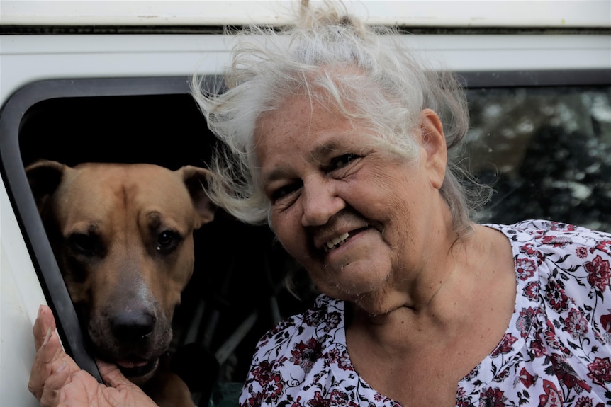 An elderly Indigenous woman standing outside her white van with a dog poking its head out the window