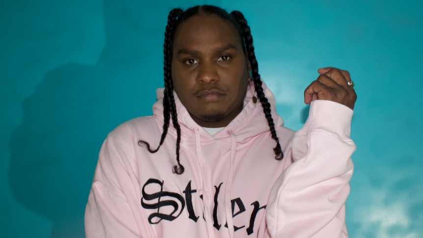 Press shot of Baker Boy; subject in pink jumper, against teal backdrop