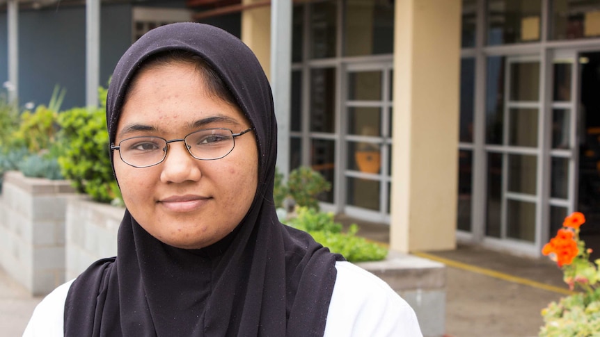 Teenage girl with glasses wearing a hijab, looking at camera with outside of school in background