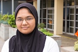 Teenage girl with glasses wearing a hijab, looking at camera with outside of school in background