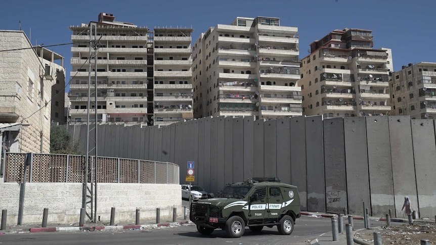 A border police car outside tall, concrete separation barrier in East Jerusalem. Apartment blocks line the land behind it.