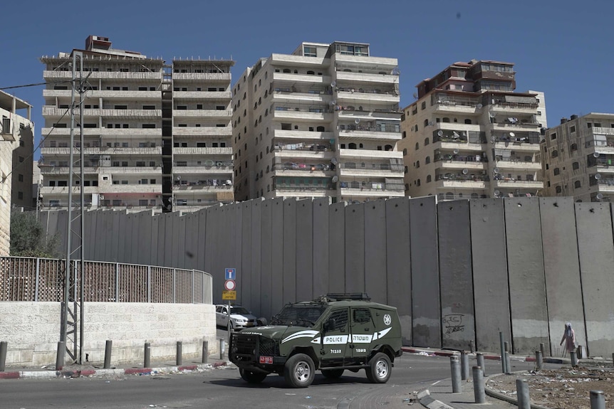 A border police car outside tall, concrete separation barrier in East Jerusalem. Apartment blocks line the land behind it.