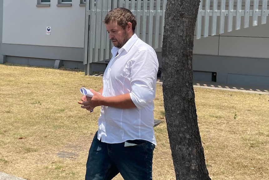 A man in a white button-up shirt walking with a tree and building behind him.