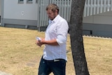 A man in a white button-up shirt walking with a tree and building behind him.