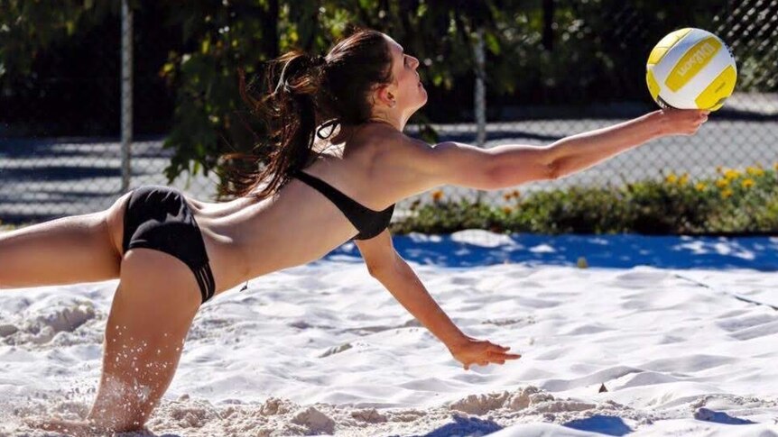 Woman with long brown hair and wearing a black bikini dives for the ball in a beach volleyball game.