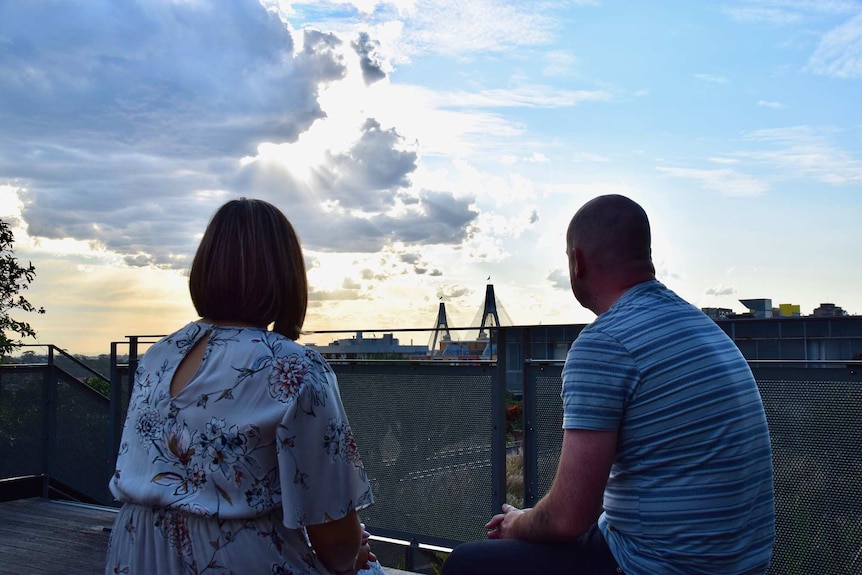 A woman and a man have their backs to the camera as they look over a city scape at sunset, remembering their stillborn son.