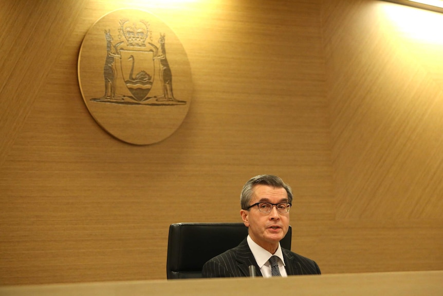 Tony Power sits at the front of the hearing room with a coat of arms on the wall behind him.