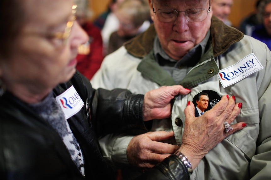 Carol Barry helps pin a button on the jacket of friend Robert Wilson before a campaign rally with Mitt Romney