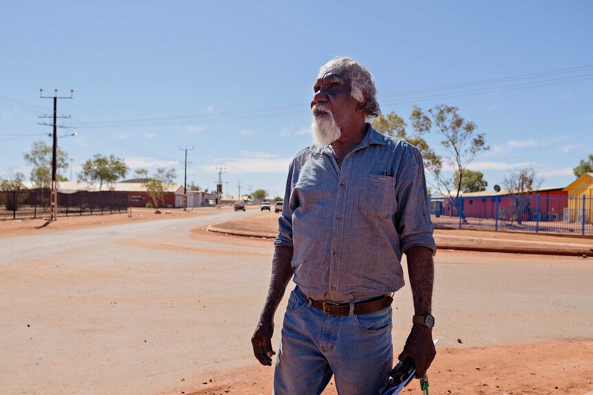 Warlpiri elder Eddie Robertson stands in a Yuendumu street looking across the camera.