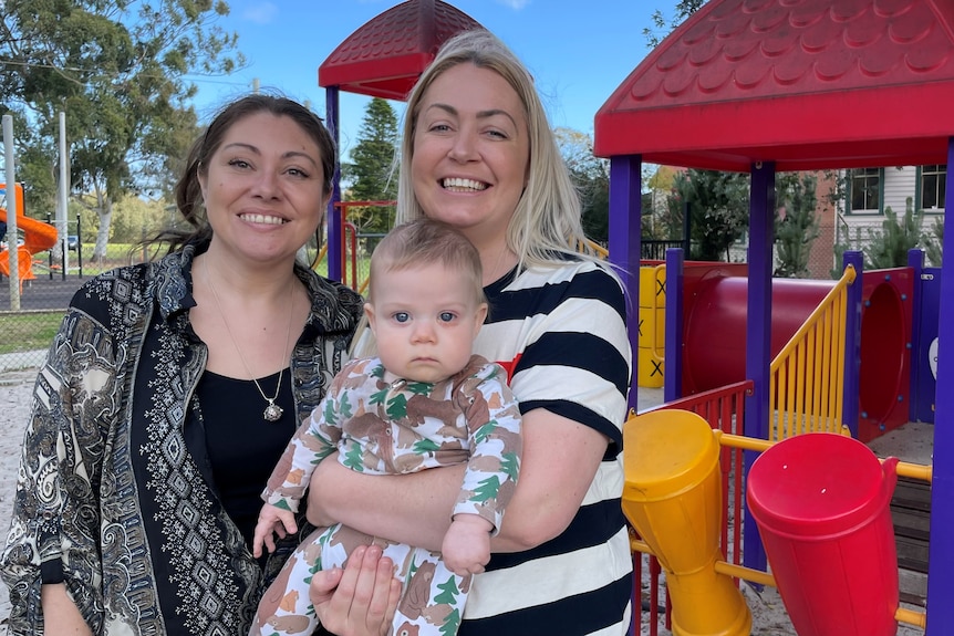 Two women stand in front of play equipment with one of them holding a baby.