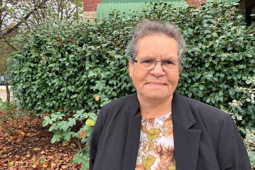 An Indigenous woman wearing glasses and a black jackets stands in front of a green bush.