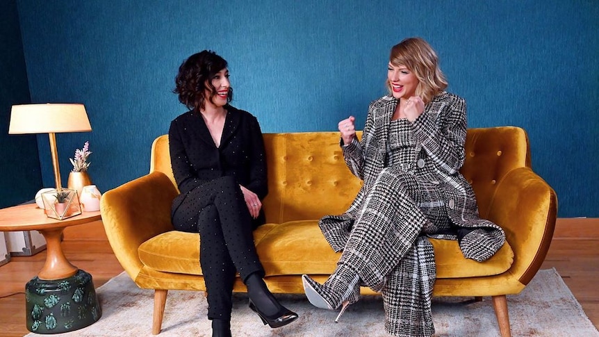 A brown haired woman and a blonde haired woman sit on a mustard couch, laughing.