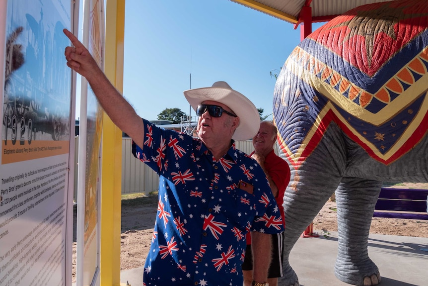 A man in an Australian flag shirt stands in front of a colourful elephant statue and points at an information board.