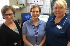 Three women smiling standing together in an office with a computer and notice board behind them