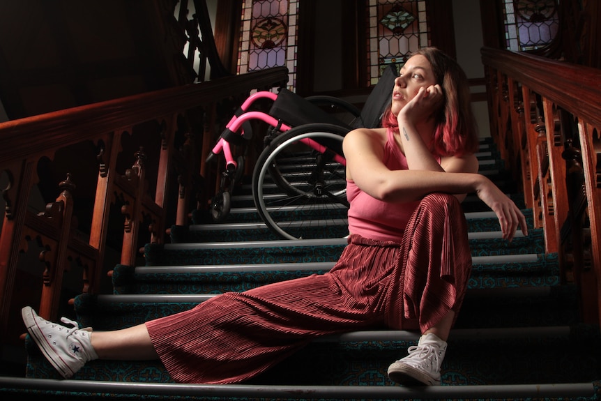 A person sits on a staircase with a pink wheelchair behind them