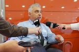 A seated man with grey hair, wearing a collared shirt and tie, with reporters holding microphones towards him.
