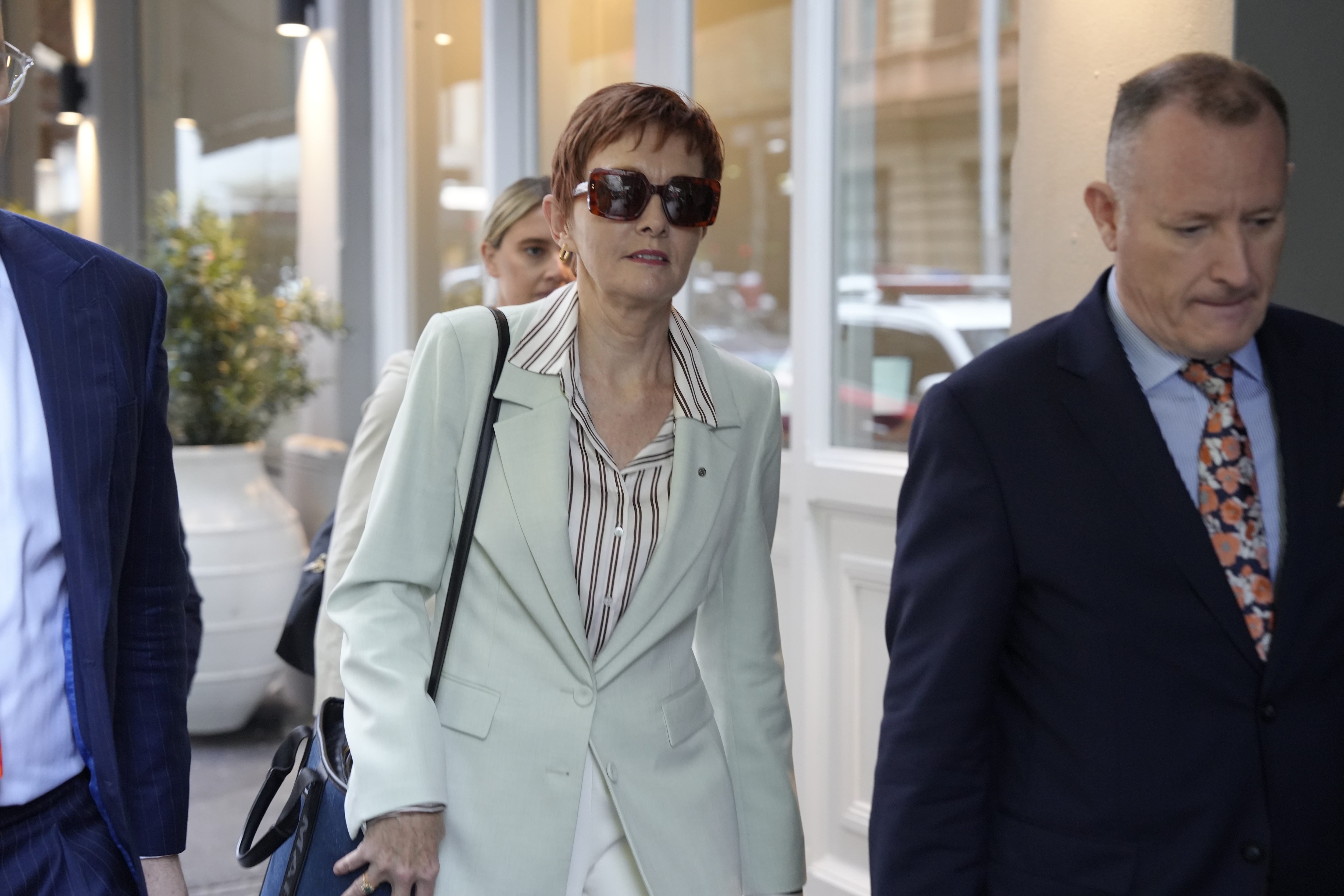 Gail wears a pale blazer and striped shirt and dark glasses. She walks alongside two men in suits.