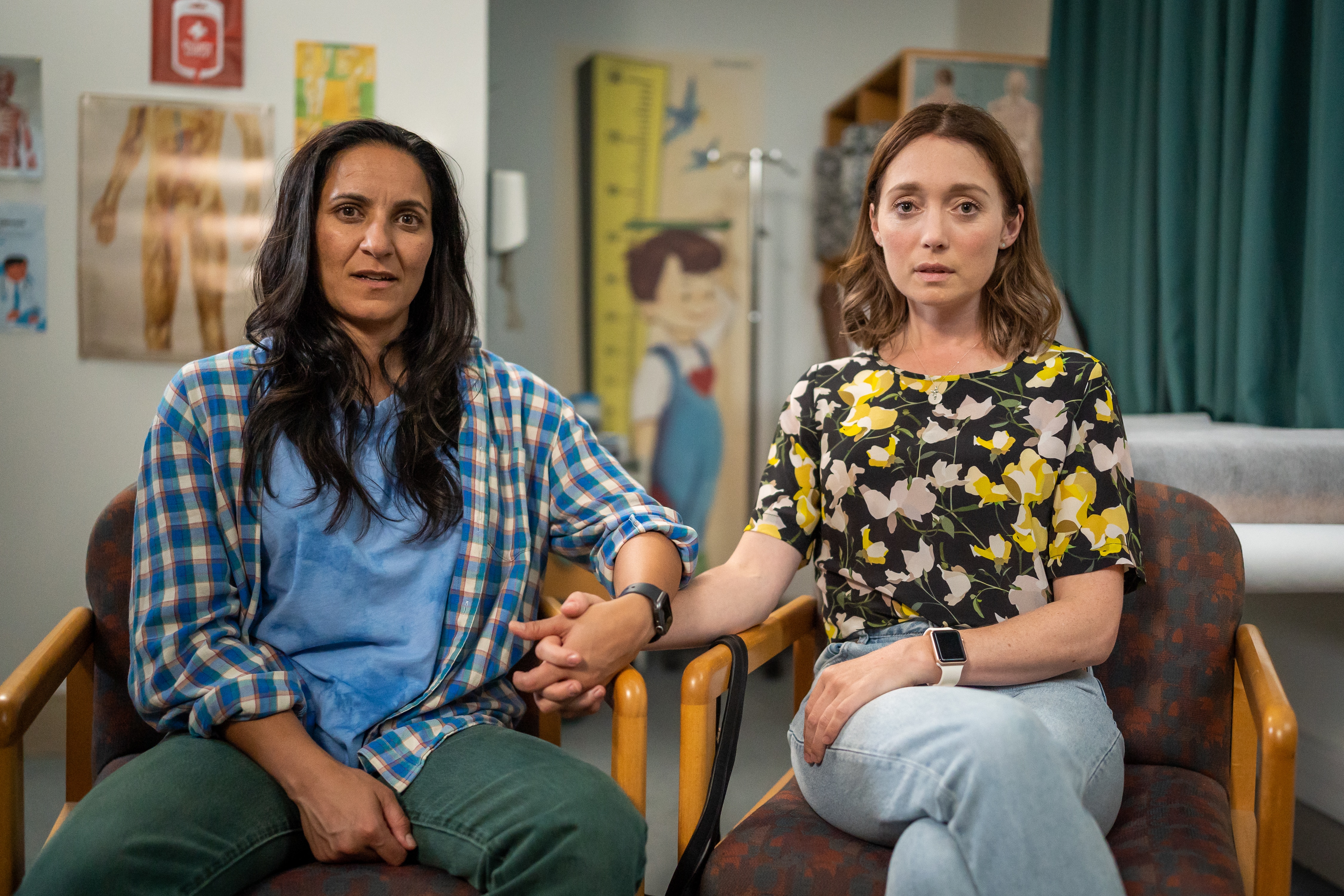 Two women hold hands looking concerned, in the chairs of a doctor's office.
