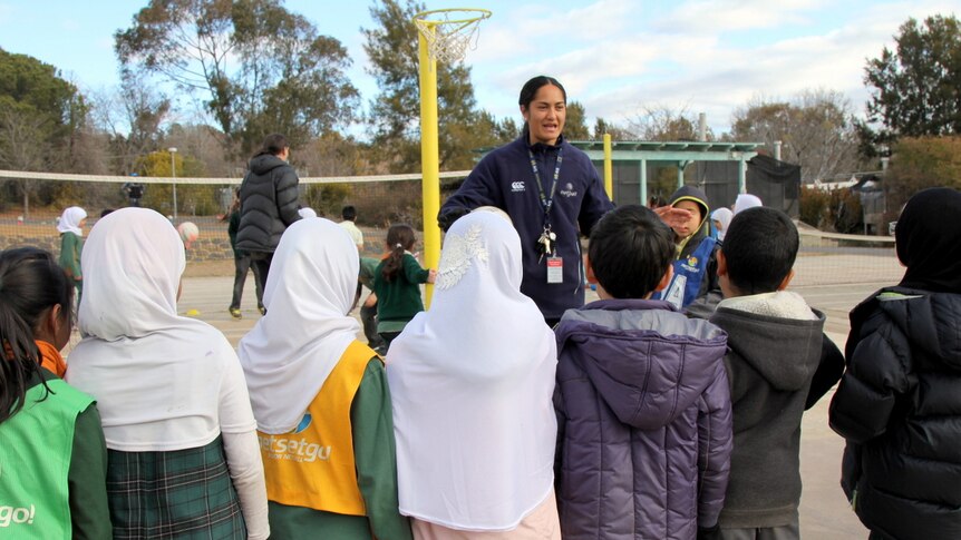 Primary school kids stand on a netball court listening to a netball player giving instructions.