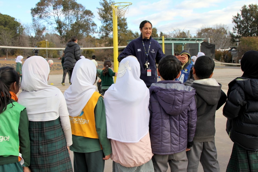 Primary school kids stand on a netball court listening to a netball player giving instructions.