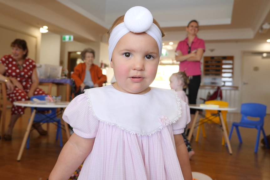 A toddler with a pink outfit and white headband looks at the camera.