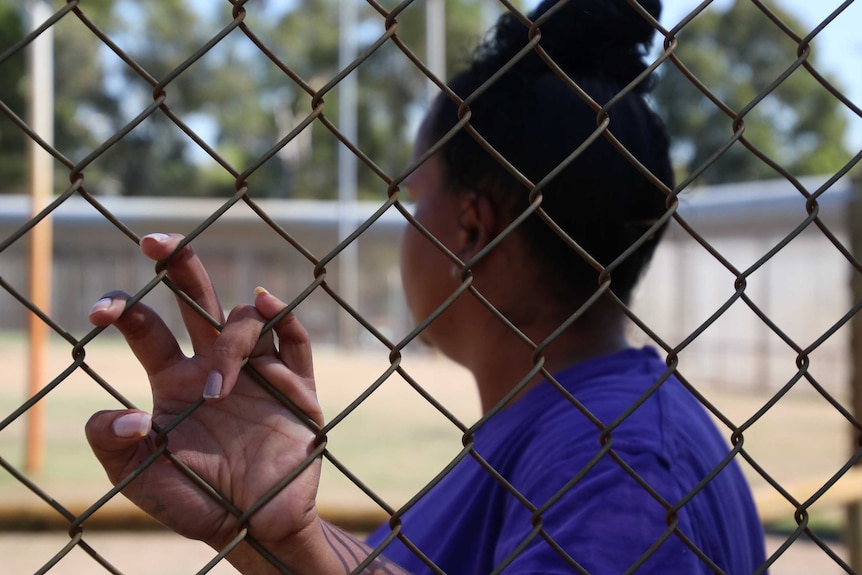 A prisoner looks into the distance as she has one hand on a prison fence.