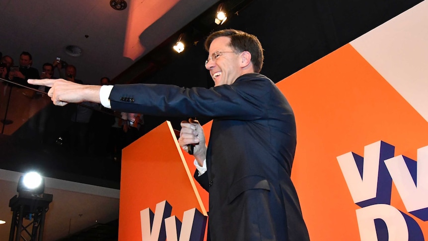 Dutch PM Mark Rutter smiles and points into the audience (out of frame) in front of "VVD" placards