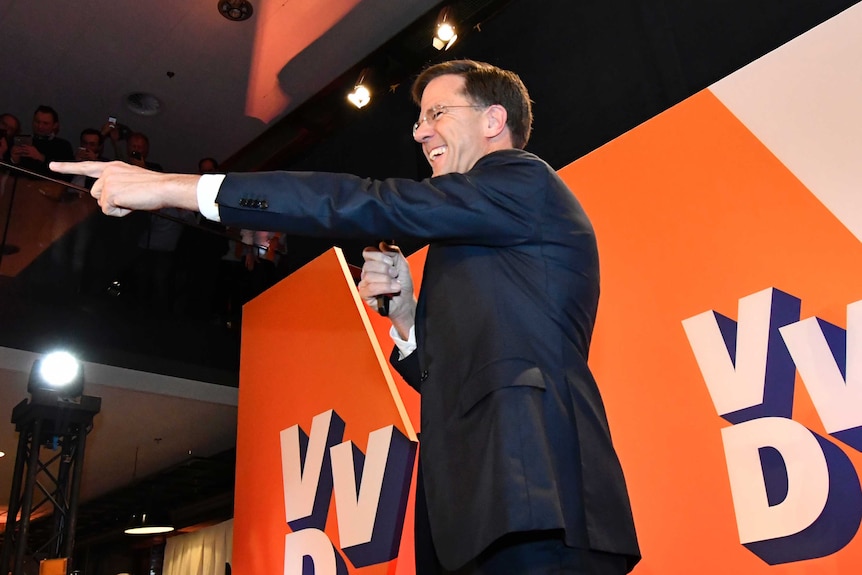 Dutch PM Mark Rutter smiles and points into the audience (out of frame) in front of "VVD" placards