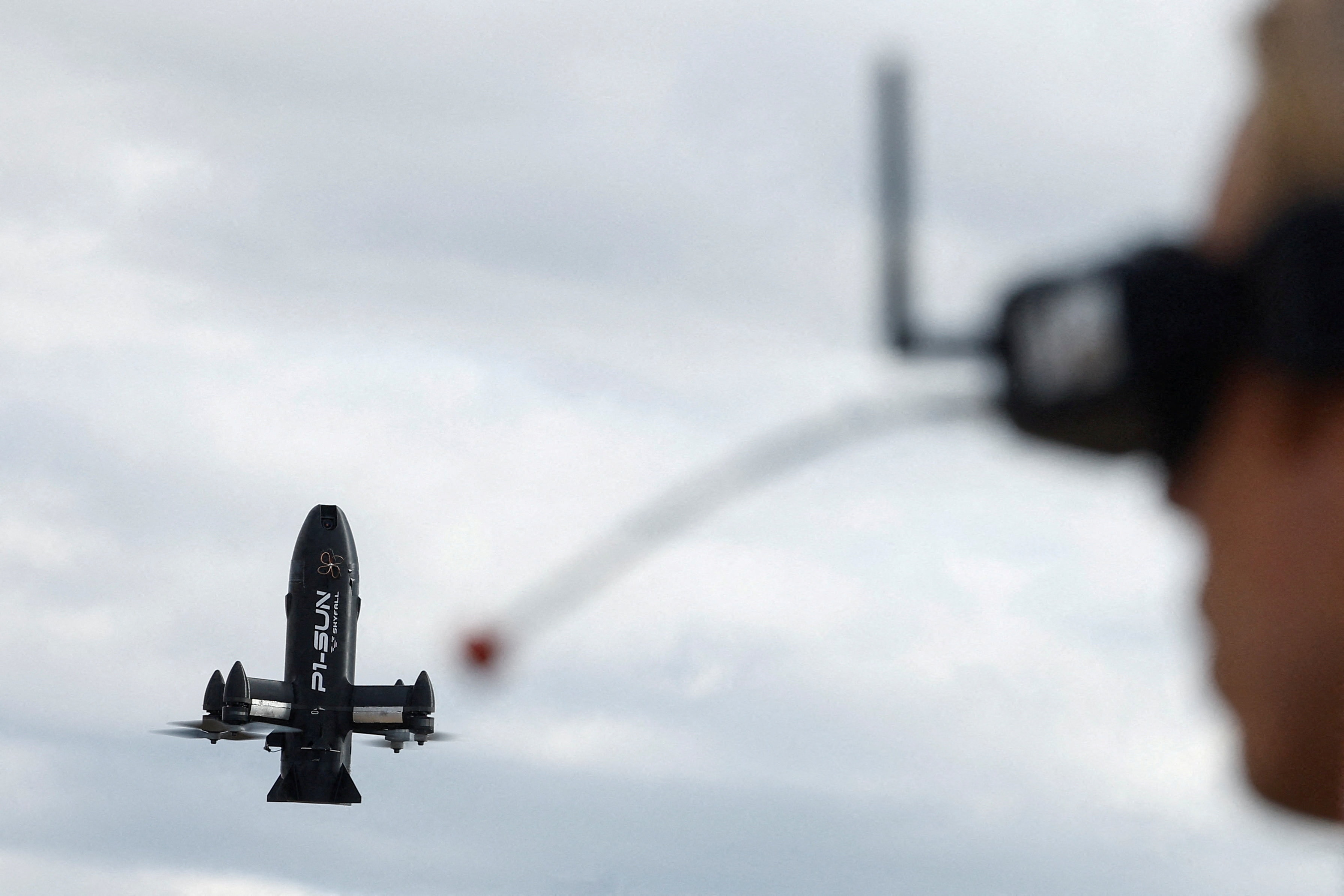 Back of man's head in foreground wearing goggles to operate a small rocket-looking drone in front of him.