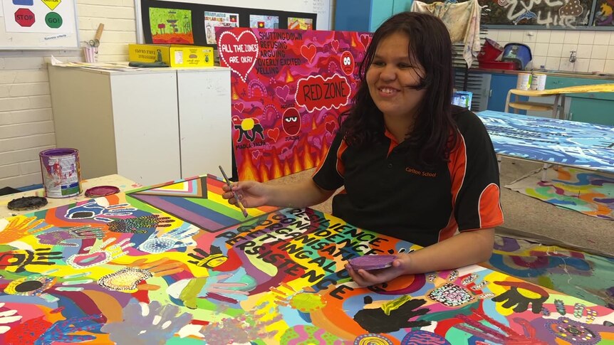 A young student in an art classroom sits at a table covered in a brightly painted mural. 