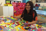 A young student in an art classroom sits at a table covered in a brightly painted mural. 