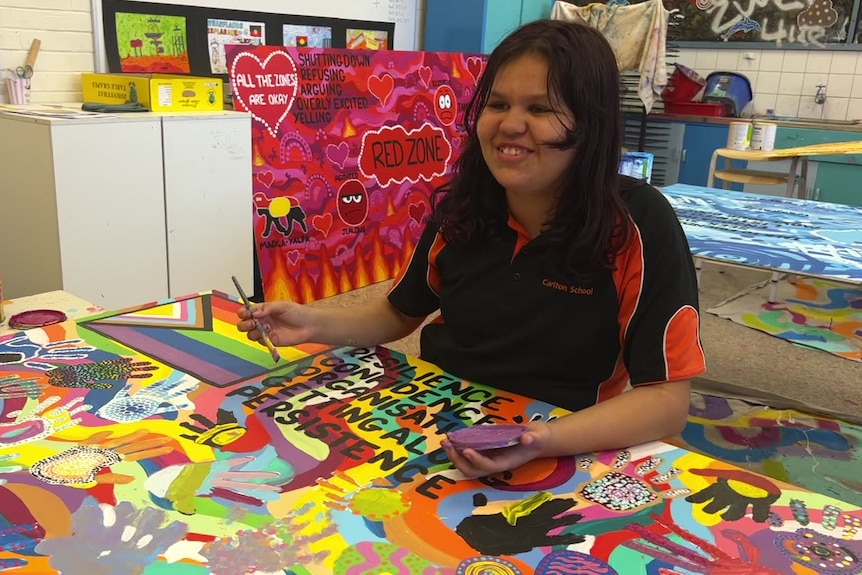 A young student in an art classroom sits at a table covered in a brightly-painted mural. 